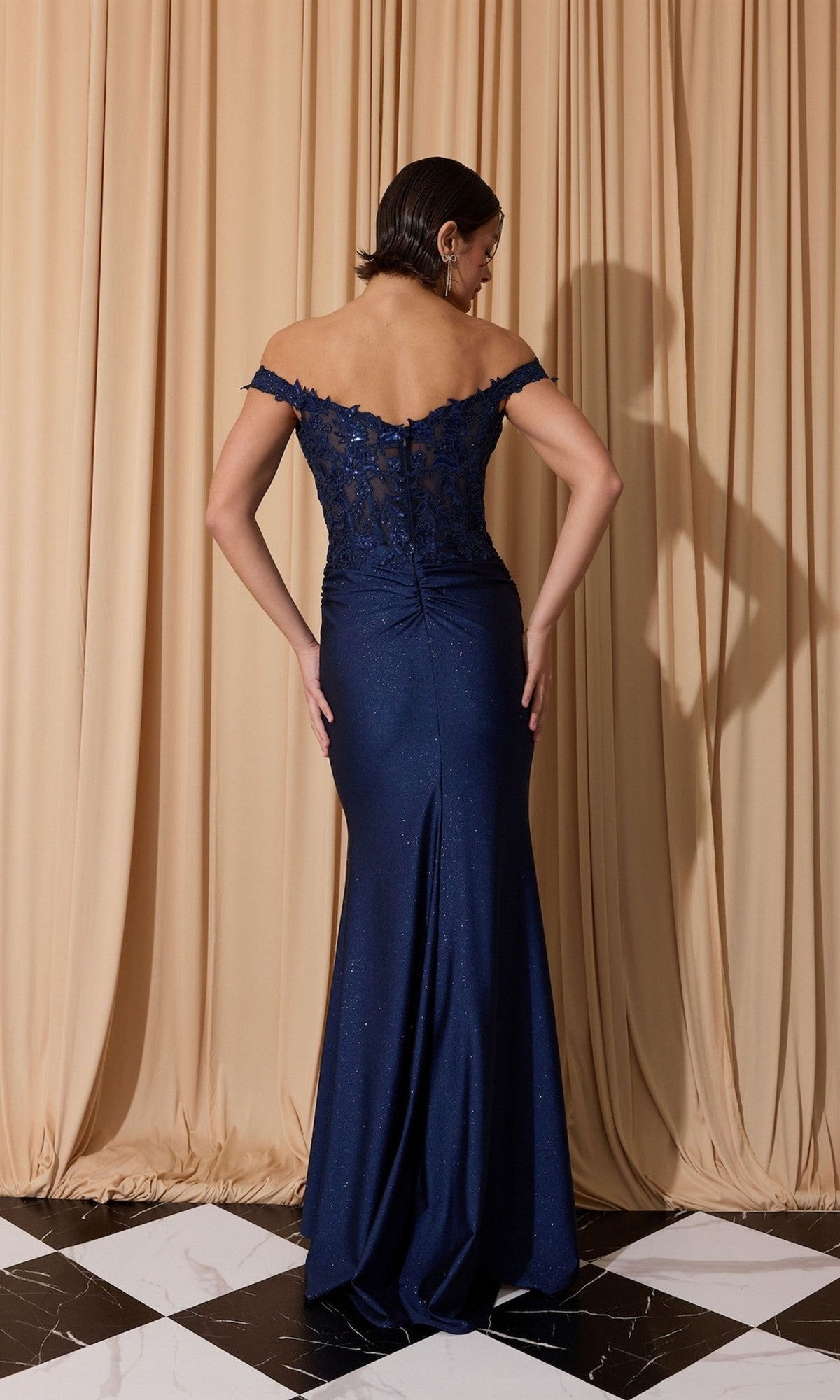 A woman stands with her back to the camera in front of beige curtains, wearing the Eureka EK173 Long Prom Dress—a navy blue off-the-shoulder gown with lace detailing—on a black and white floor.